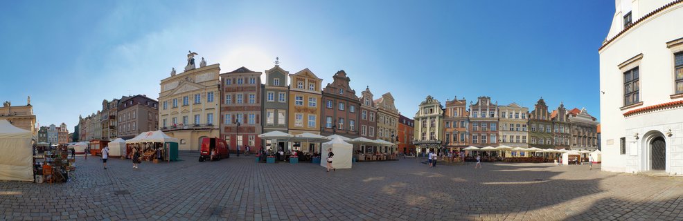 Wide panoramic view of Poznań Old Market Square showing colorful historic buildings, market tents, cobblestone plaza, and pedestrians under blue sky