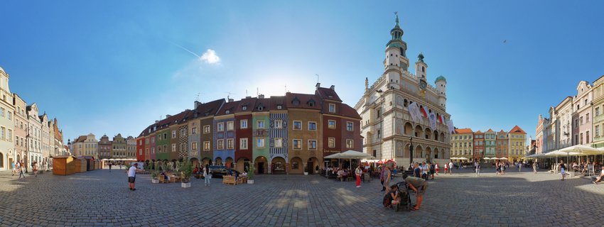 Panoramic view of Poznań Old Market Square showing colorful historic buildings and the ornate Town Hall under blue sky with people walking on cobblestone pavement
