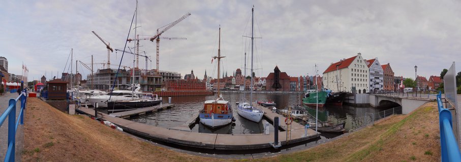 Wide harbor view of Gdansk waterfront showing docked boats, historic red-roof granary buildings, medieval wooden port crane, construction cranes, and bridge along Motlawa River under overcast sky