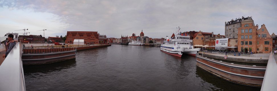 Wide waterfront view of Gdańsk's Motława River showing red brick historic buildings, white ferry boats docked at wooden piers, people walking on promenades, and overcast sky
