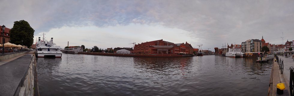 Wide waterfront view of Gdansk's historic Old Town showing red brick buildings along the Motława River with docked white ships and stone walkways on both sides under cloudy sky