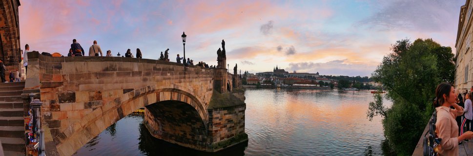 Stone arch bridge with tourists and baroque statues spanning a river at sunset, with castle complex visible on distant hill under pink and orange sky