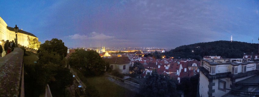 Panoramic evening view of Prague showing illuminated Prague Castle on the left, Petřín Tower on a forested hill to the right, and red-roofed historic buildings with city lights under a deep blue twilight sky