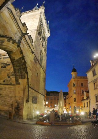 Illuminated Gothic stone tower with arched gateway at night, outdoor café seating with umbrellas on cobblestone plaza, deep blue twilight sky, Prague