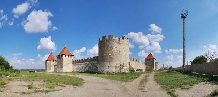 Medieval stone fortress with multiple towers featuring red-tiled roofs under a blue sky with white clouds, surrounded by green grass and dirt paths