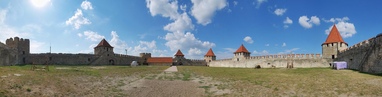 Panoramic view of medieval stone fortress with multiple towers featuring red-tiled roofs surrounding a grassy courtyard under blue sky with white clouds