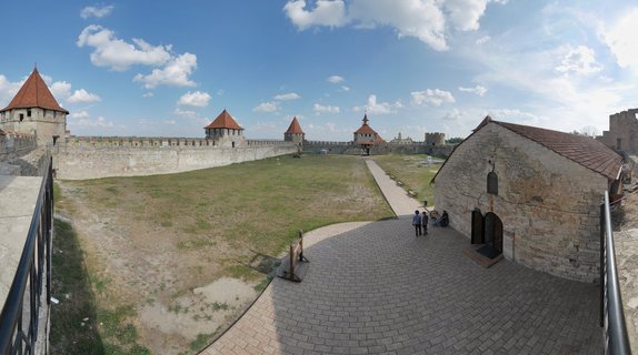 Wide view of Bender Fortress courtyard showing medieval stone walls, multiple towers with red conical roofs, paved walkways, green grass, and visitors walking near stone buildings under blue sky with white clouds