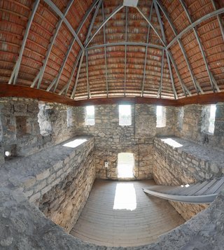 Interior of a circular stone tower with beige limestone walls, wooden floor, multiple window openings with bright daylight, and a conical roof with red tiles and metal support beams