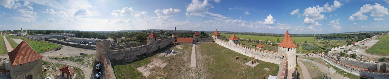 Panoramic aerial view of Bender Fortress showing medieval stone walls, multiple towers with red conical roofs, green grassy courtyards, and blue sky with white clouds