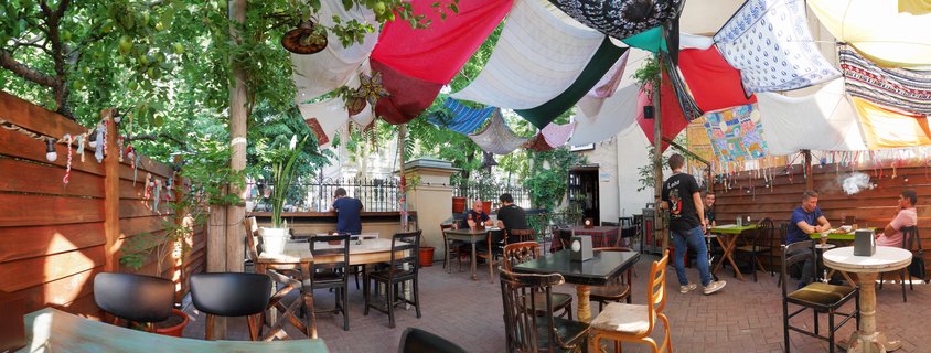 Outdoor café patio with colorful fabric canopies hanging overhead, wooden tables and chairs, people dining, surrounded by trees and wooden fencing