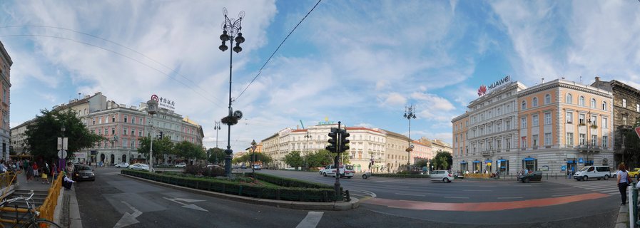 Panoramic street view of Budapest showing historic multi-story buildings with ornate facades, traffic intersection, street lamps, and pedestrians under blue sky with clouds