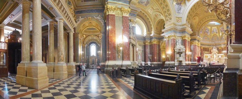 Interior view of an ornate cathedral with marble columns, gold decorations, vaulted ceilings with paintings, checkered floor, and wooden pews