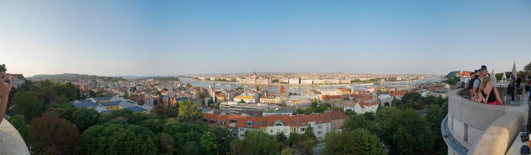 Panoramic cityscape of Budapest showing the Danube River, Parliament Building, and dense urban architecture with red-tiled roofs viewed from an elevated stone terrace with tourists