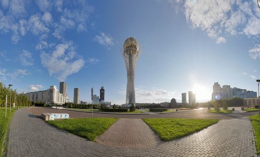 White lattice tower with golden sphere at top in center of wide city plaza with paved walkways, green lawns, and modern buildings under blue sky with sun flare