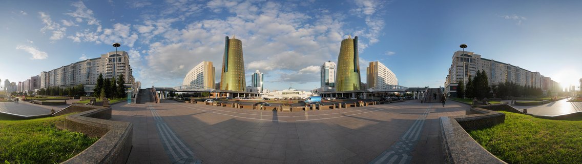 Panoramic view of two golden cylindrical tower buildings in a modern plaza with paved walkways, green lawns, and blue sky with clouds in Astana, Kazakhstan