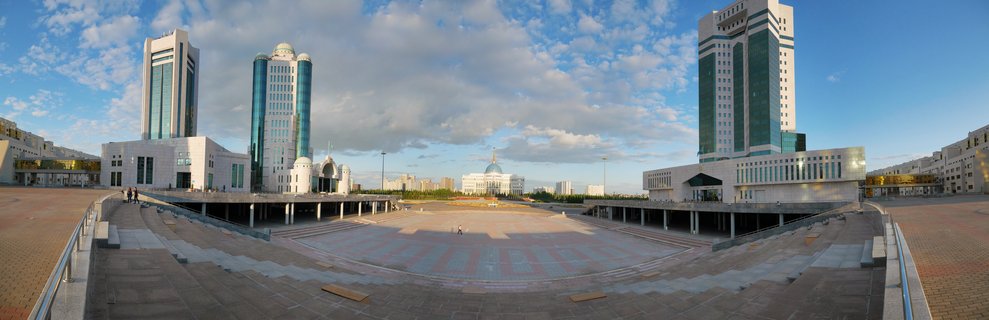 Wide panoramic view of the Ak Orda Presidential Palace with blue dome centered between tall modern glass office towers, overlooking a large paved public plaza under blue sky with scattered clouds in Astana, Kazakhstan
