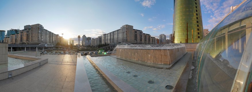 Urban plaza with water fountains in foreground, golden cylindrical tower on right, apartment buildings and sunset in background, Astana Kazakhstan