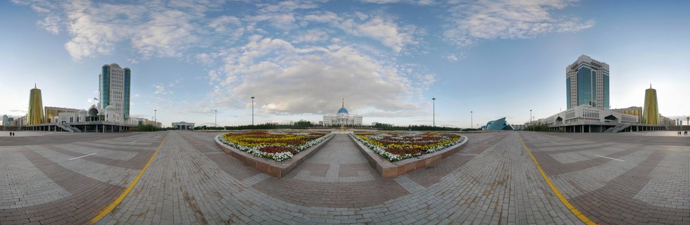 Wide panoramic view of Astana's central square with the blue-domed Presidential Palace in the center, modern skyscrapers on both sides, colorful flower beds in the foreground, and blue sky with scattered clouds