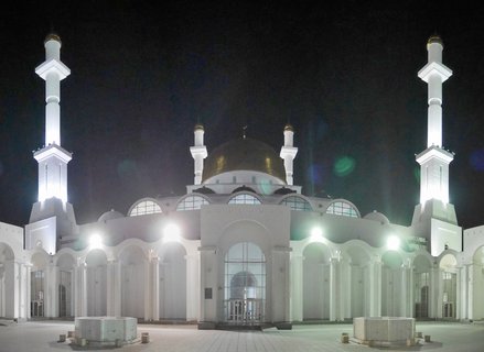 Night photograph of Hazret Sultan Mosque in Astana showing white building with golden central dome, four illuminated minarets, arched entrance, and paved courtyard with fountains
