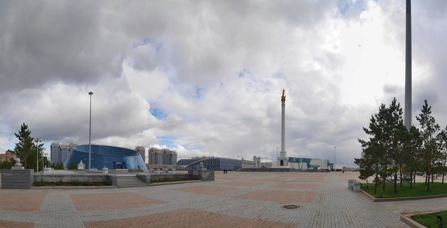 Wide city plaza in Astana with tall white monument column, modern blue glass buildings, paved walkway, evergreen trees, and cloudy sky