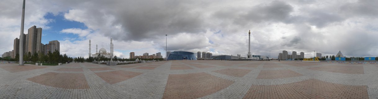 Wide panoramic view of a large paved city square in Astana with a white mosque featuring multiple minarets on the left, a tall monument column on the right, and modern apartment buildings in the background under cloudy skies