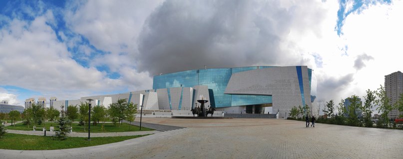 Wide exterior view of the National Museum of Kazakhstan showing a large modern building with blue glass and white angular stone walls, a paved plaza with visitors, green landscaping with young trees, and dramatic cloudy sky overhead