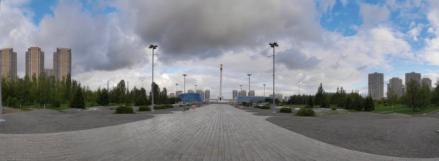 A wide view of a paved public plaza in Astana, Kazakhstan, featuring a central white monument with golden ornament, flanked by modern high-rise buildings and landscaped greenery under a cloudy sky.