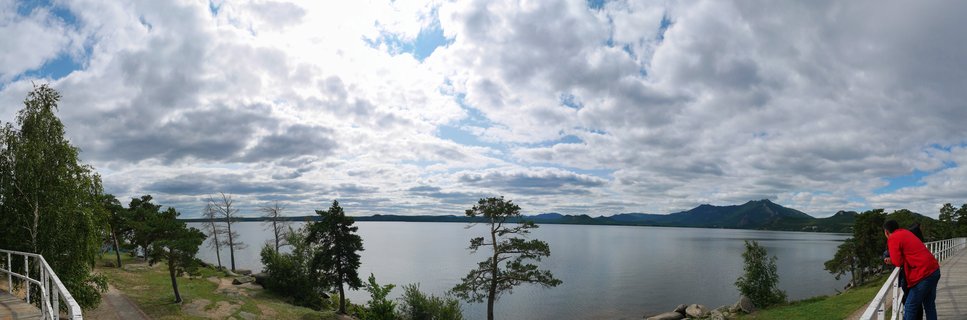 Panoramic landscape showing Lake Burabay in Kazakhstan with calm water, forested shoreline with birch and pine trees, distant mountains on the horizon, dramatic cloudy sky, and a person in red jacket at a white-railed viewpoint on the right