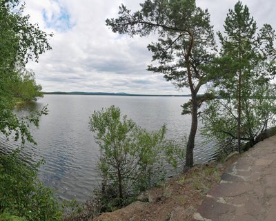 A lakeside view showing calm water stretching to distant hills, with pine trees on the right, deciduous trees on the left, and a stone pathway along the shore in the foreground