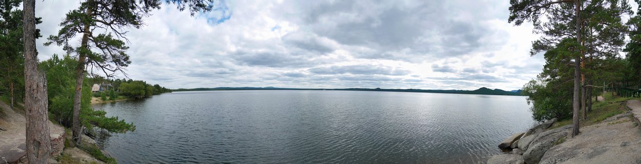 Wide panoramic photograph of Lake Burabay showing calm blue-grey water with pine trees on both sides of the frame, rocky shoreline, and cloudy sky with distant hills