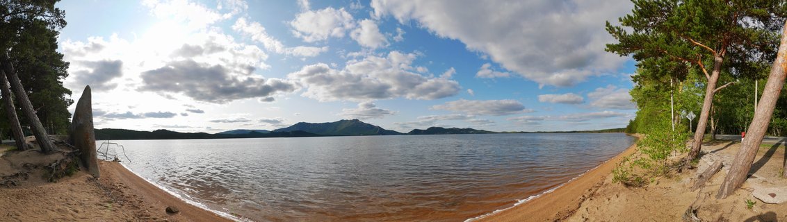 Wide panoramic photograph of Lake Burabay showing sandy beach, calm brown water, pine trees on both shores, and green mountains in the distance under blue sky with white clouds