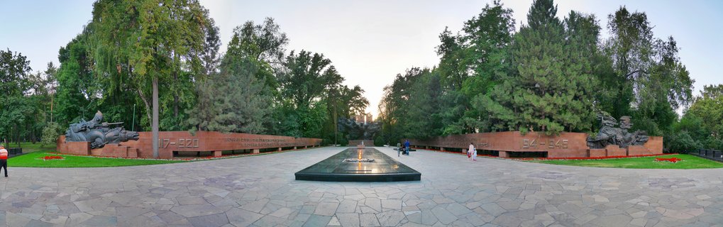 Memorial complex featuring bronze sculptures of Soviet WWII soldiers in front of red granite walls with inscriptions, centered around a fountain water feature in Panfilov Park, Almaty, Kazakhstan.