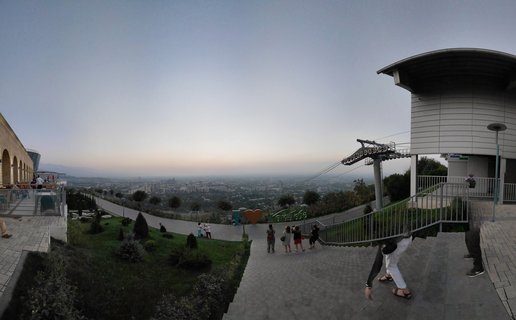 Panoramic view from Kok Tobe Hill showing cable car station, walking visitors, heart sculpture, and Almaty cityscape at twilight in Kazakhstan