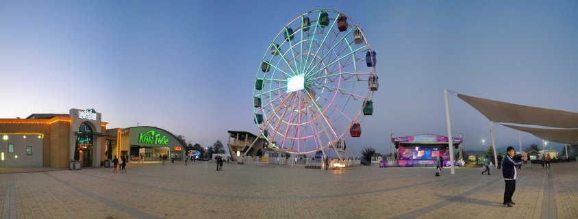 Large illuminated Ferris wheel with multi-colored lights at an amusement park plaza during twilight, with people walking on brick pavement and buildings with signage visible