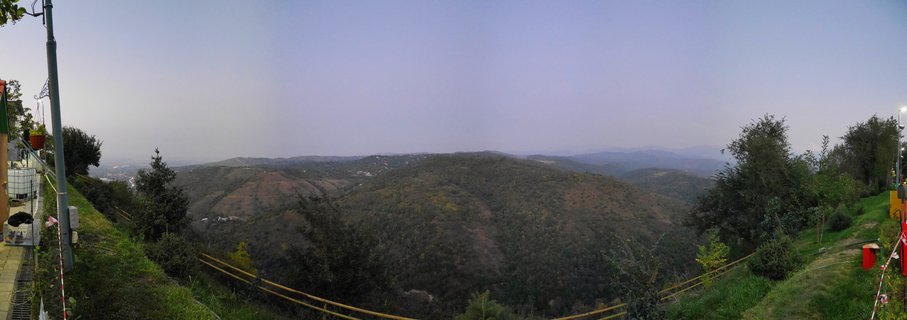 Wide panoramic photograph of forested mountain hills near Almaty, Kazakhstan at dusk with yellow safety railings in the foreground and hazy distant mountain ranges under a pale blue evening sky