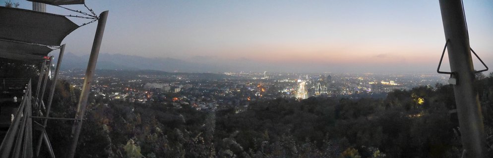 Panoramic view of Almaty city lights at twilight with mountains in the background and dark hillside vegetation in foreground