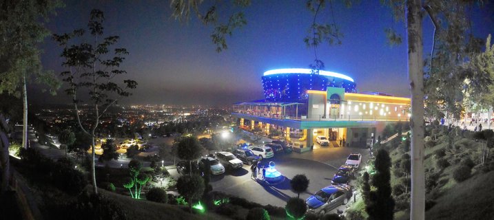 Nighttime panoramic view of a brightly lit modern building with blue LED roof lighting and a parking lot, overlooking the illuminated cityscape of Almaty, Kazakhstan with trees in the foreground