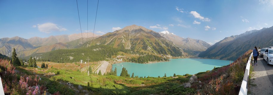 Panoramic view of turquoise Big Almaty Lake surrounded by mountain ranges with coniferous forests, cable car lines, and a viewing platform with visitors on the right side under blue sky