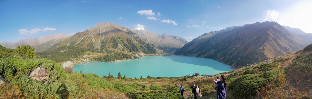 Panoramic view of Big Almaty Lake showing turquoise alpine waters surrounded by brown and green mountain slopes under a blue sky with scattered clouds, with tourists visible in the foreground