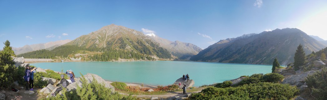 Panoramic landscape showing turquoise Big Almaty Lake surrounded by mountain peaks in the Tian Shan range near Almaty, Kazakhstan with tourists on rocky outcrops among coniferous trees under clear blue sky