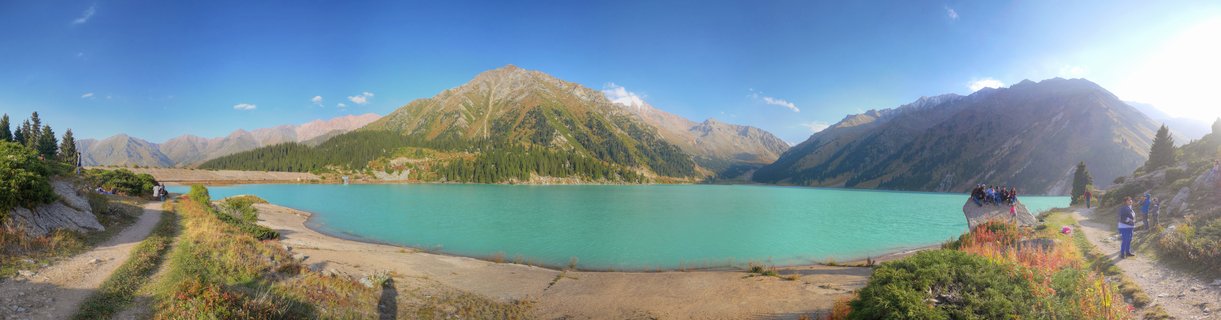 Panoramic view of Big Almaty Lake in Kazakhstan showing turquoise glacial alpine lake surrounded by Tien Shan mountain peaks with coniferous forest slopes, hiking trails with tourists on both shores, clear blue sky, and golden hour sunlight during late afternoon in early autumn