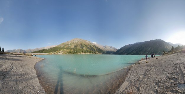 Panoramic view of turquoise alpine lake surrounded by mountains with coniferous forests and visitors on the rocky shoreline under clear blue sky