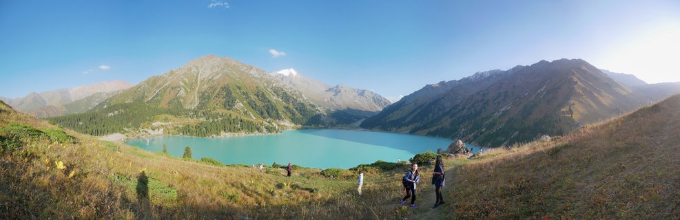 Panoramic view of turquoise Big Almaty Lake surrounded by mountains in Kazakhstan, with hikers walking on grassy hillside in foreground under clear blue sky