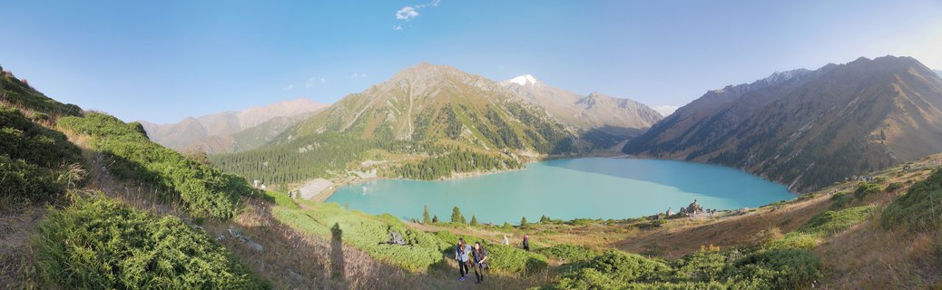Wide panoramic landscape showing turquoise alpine lake surrounded by brown and green mountains under clear blue sky with hikers on grassy foreground slope