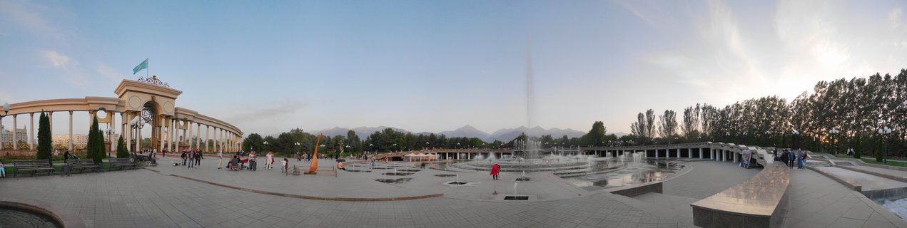 Wide panoramic photograph of a large public square in Almaty with a columned government building flying the Kazakhstan flag, multiple water fountains, pedestrians walking on paved plaza, trees, and mountains visible in the distance under a clear sky