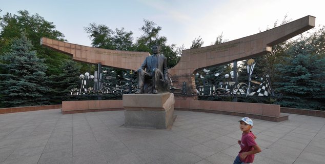 Bronze statue of seated man on stone pedestal with curved memorial structure and decorative metalwork in paved plaza with trees
