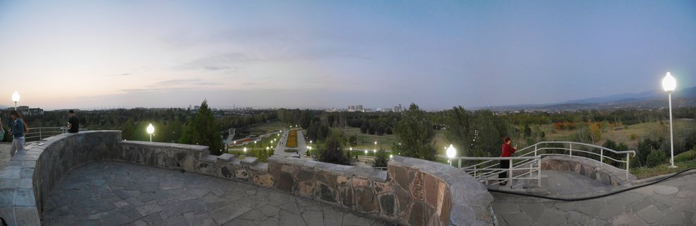 Panoramic view from a stone terrace at Kok Tobe hill showing an illuminated pathway descending through trees toward Almaty city with mountains visible on the horizon during twilight