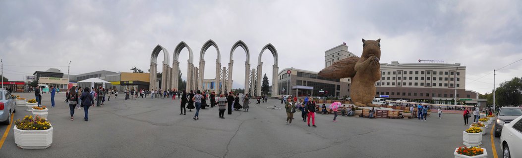 Wide panoramic street view showing five tall white arched monuments in center background, a large brown squirrel sculpture on right, pedestrians walking across paved plaza with flower planters, overcast sky, urban buildings surrounding the square