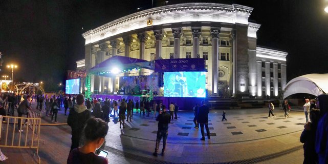 Night view of a large columned building with bright exterior lighting, outdoor concert stage with purple and blue lights, LED screens, and crowds of people in a paved plaza