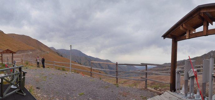 Mountain viewpoint with wooden gazebos, gravel pathway, and wooden fencing overlooking golden-brown hillsides and distant mountain ranges under cloudy sky in Kazakhstan